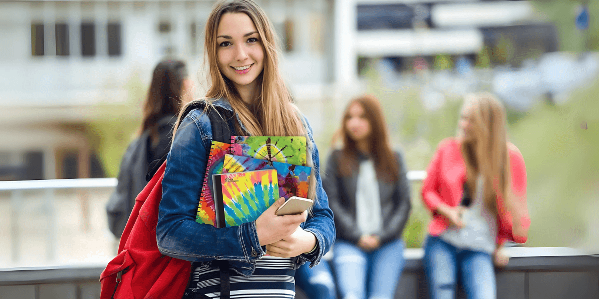 A smiling teenage girl holding colorful tie-dye notebooks and a smartphone stands in the foreground on a school campus, wearing a denim jacket and red backpack, with other casually dressed students socializing in the background.