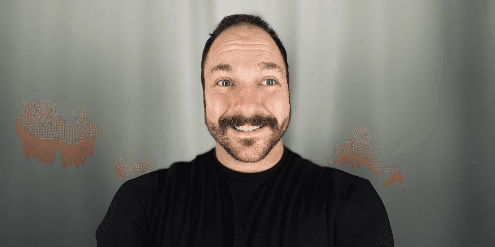 Smiling man with short dark hair and mustache wearing a black shirt, standing against a blurred gray background.
