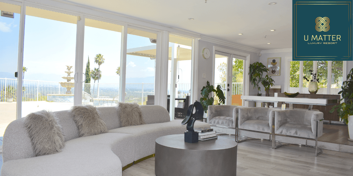 Interior of a luxury resort lounge with large windows showcasing a scenic outdoor view of a pool and fountain. A modern sofa, chairs, and decorative elements create a comfortable and elegant atmosphere, with the 'U MATTER Luxury Resort' logo in the corner.