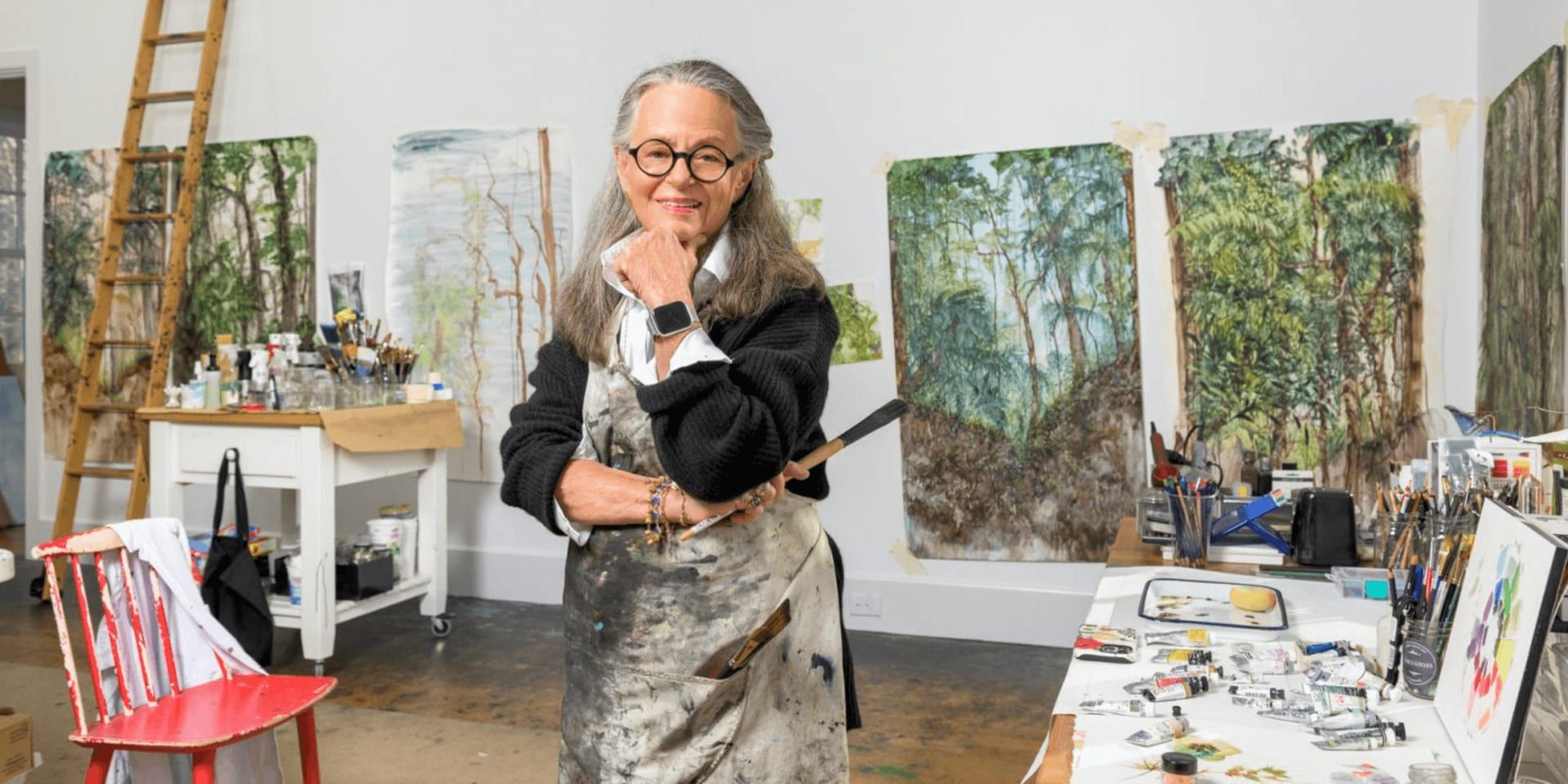 Margery Thomas-Mueller standing in her art studio surrounded by abstract landscape watercolor paintings and painting supplies.
