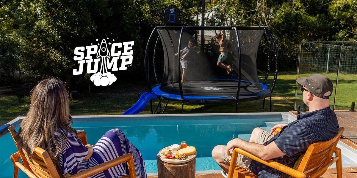Family relaxing by pool watching kids jump on a SpaceJump trampoline with safety net and slide in backyard.