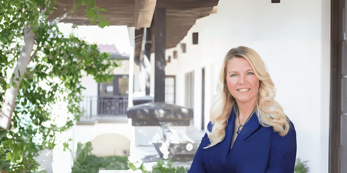 Portrait of a professional woman with long blonde hair, standing outside in front of a modern white building with greenery.