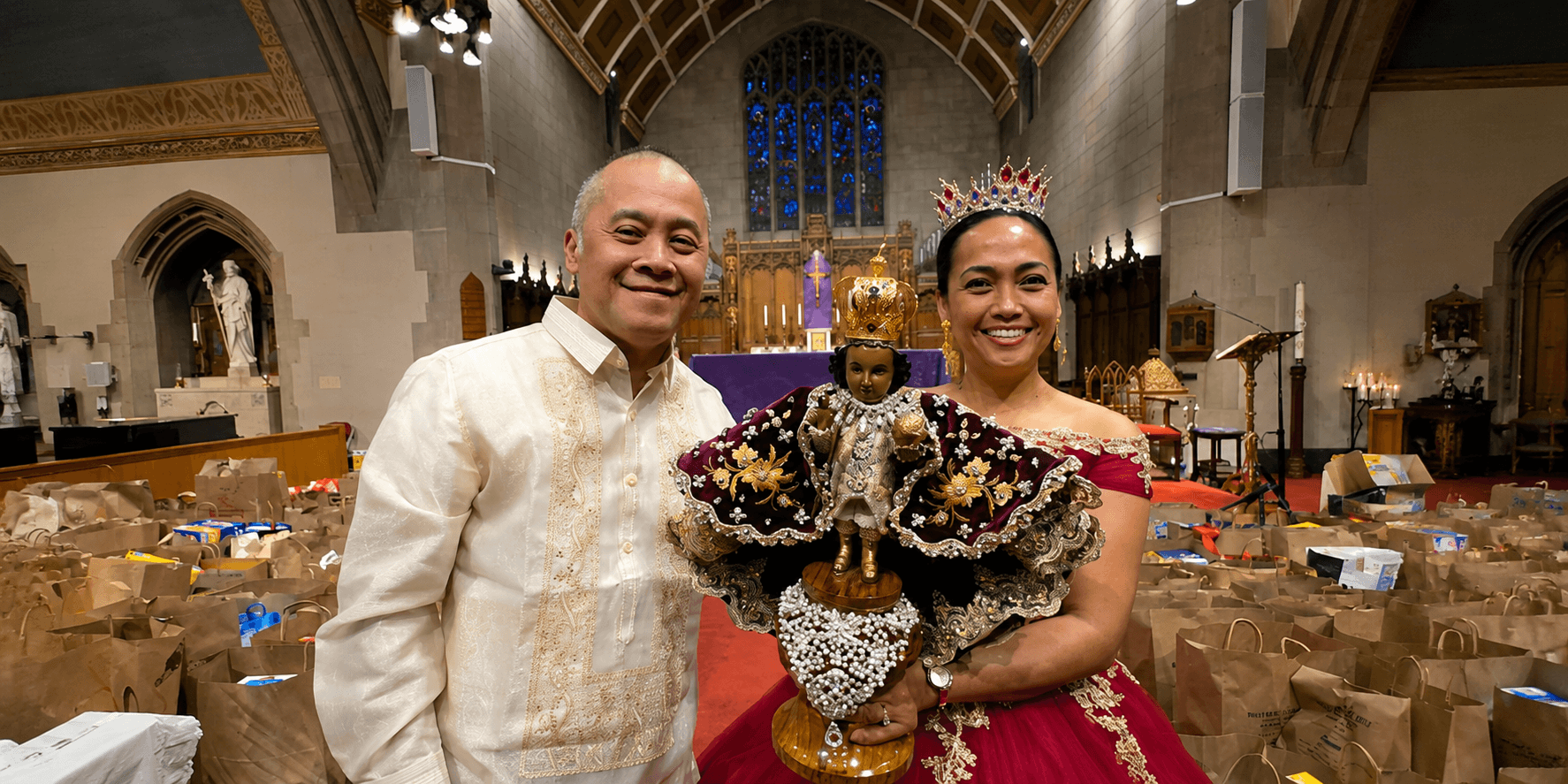 Couple holding a Santo Niño statue inside a church with donated food bags in the background.