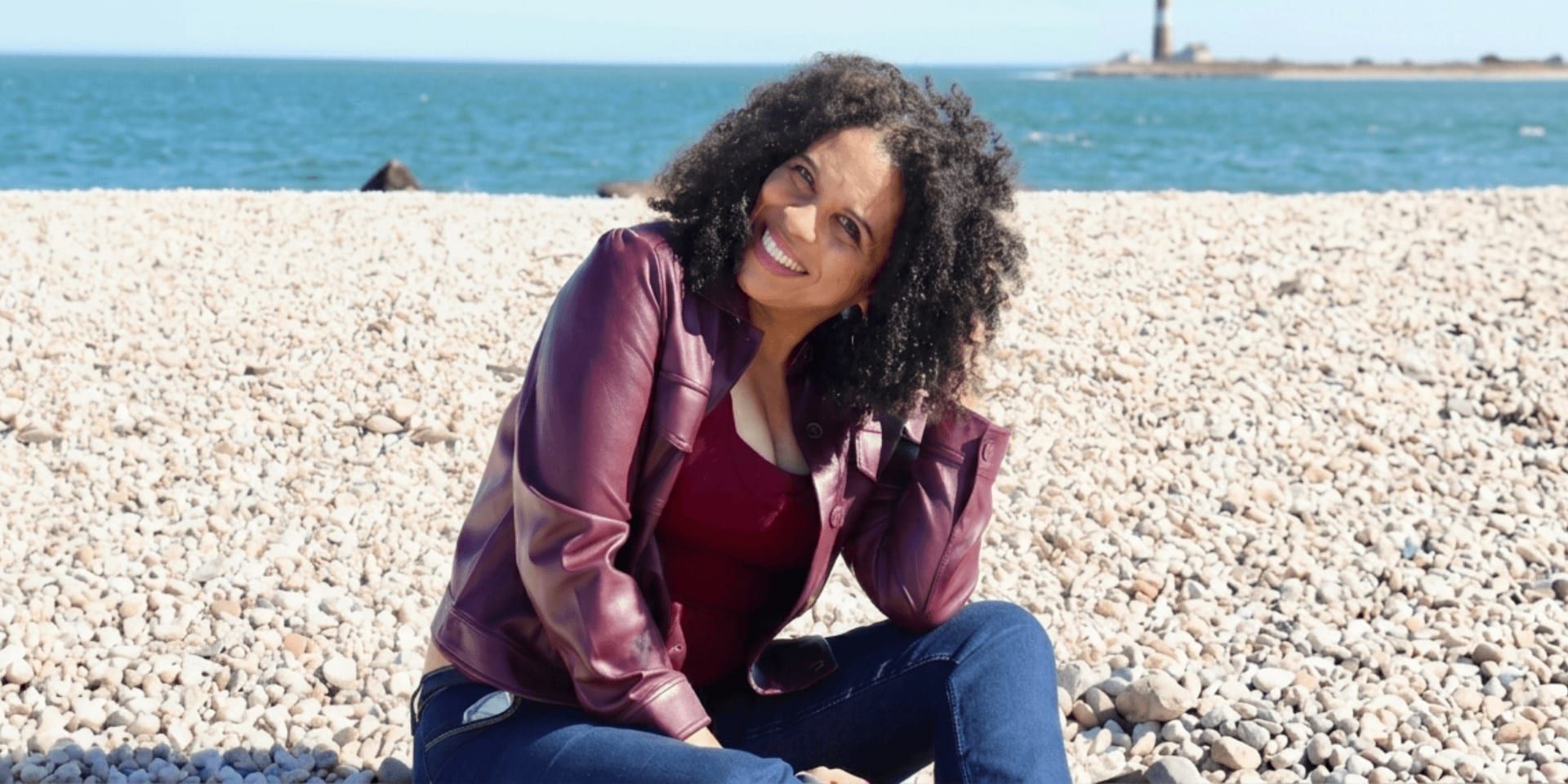 Smiling woman with curly hair sitting on a rocky beach, ocean and distant lighthouse in background.