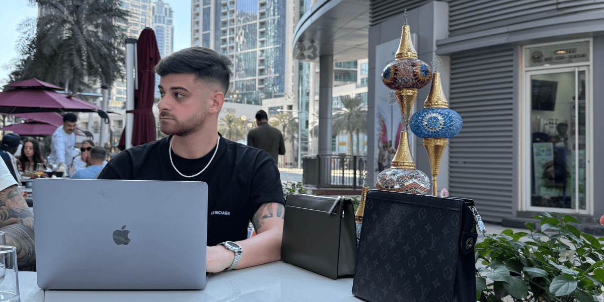 A young man with a laptop sits outdoors in Dubai, surrounded by luxury handbags, symbolizing ambition and entrepreneurship.