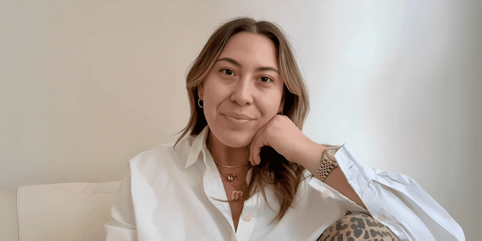 A young woman wearing a white shirt, a layered necklace, and a watch, smiling while resting her c