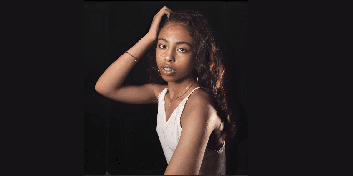 A young woman with curly hair, wearing a white top, gazing into the camera with a serene expression