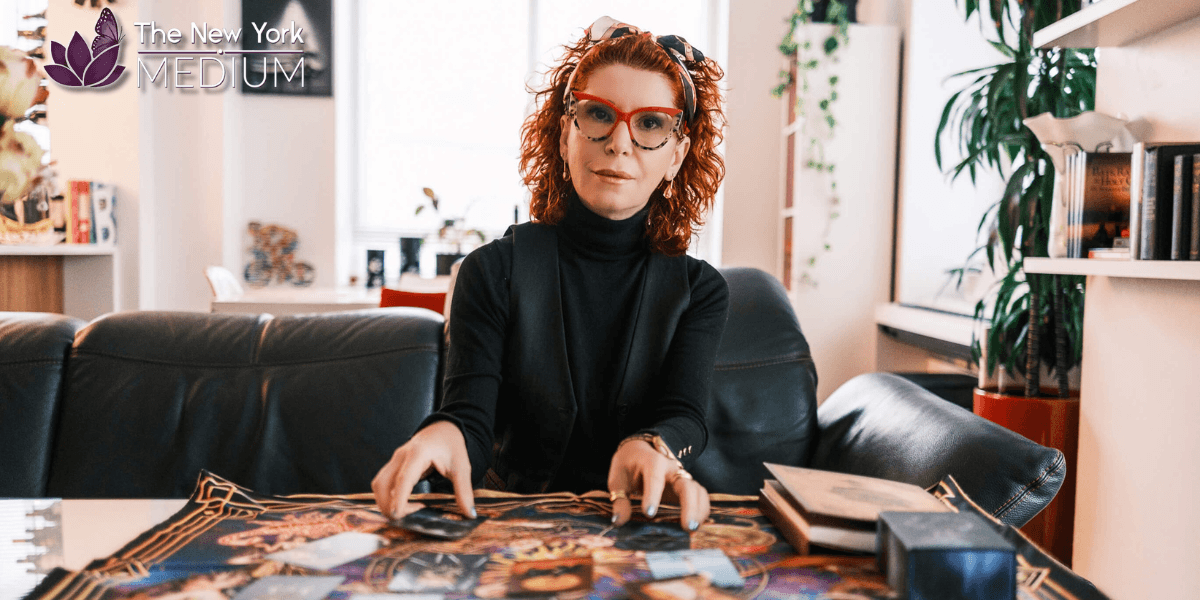 Woman with red hair and glasses performing a tarot reading at a table in a cozy, book-filled room.