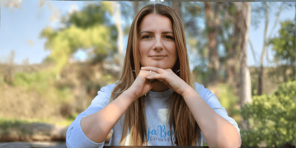A woman with long hair, wearing a sweatshirt, sits with her hands clasped, surrounded by nature, reflecting a calm demeanor.