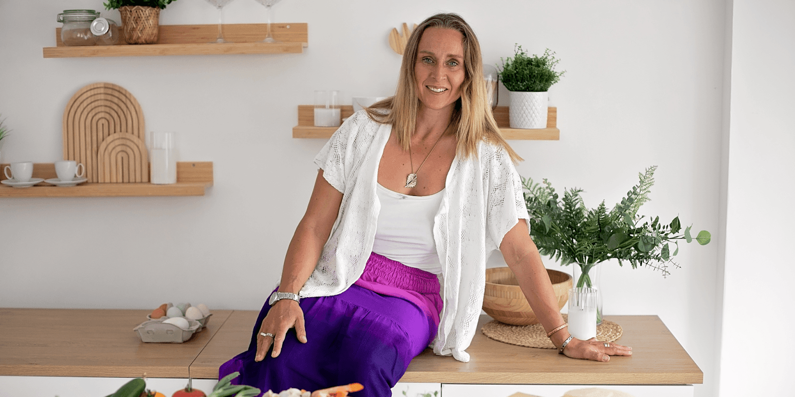 A woman with long hair, wearing a white top and purple skirt, sitting on a wooden counter surrounded by fresh vegetables and eggs in a modern, bright kitchen.