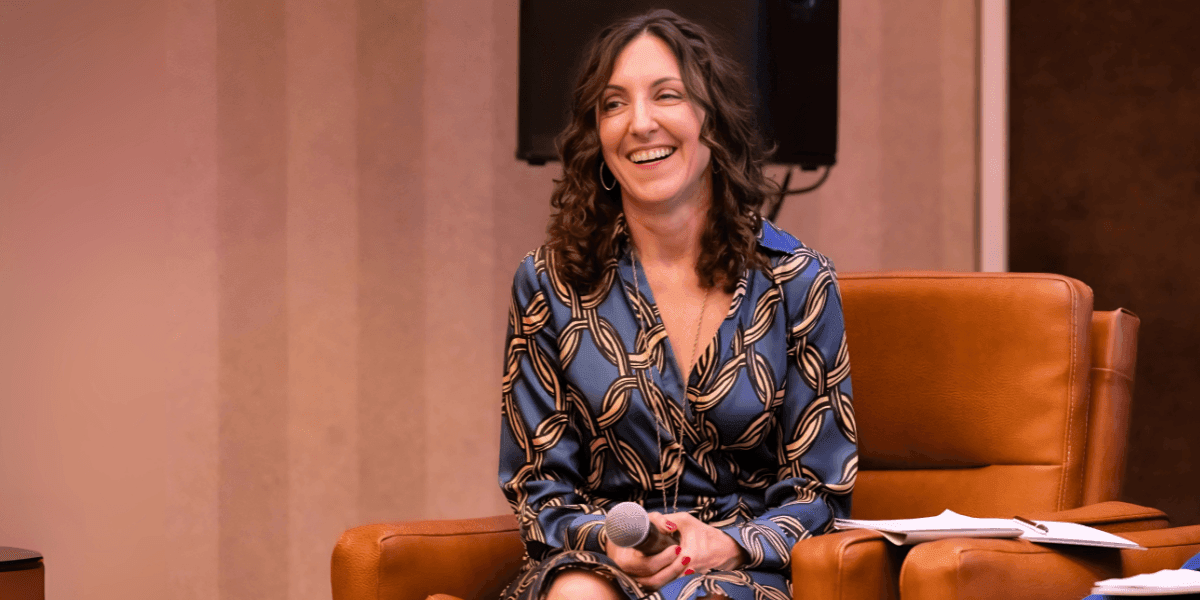 Elizabeth De Sevo smiling while holding a microphone, sitting on a leather chair at an event.