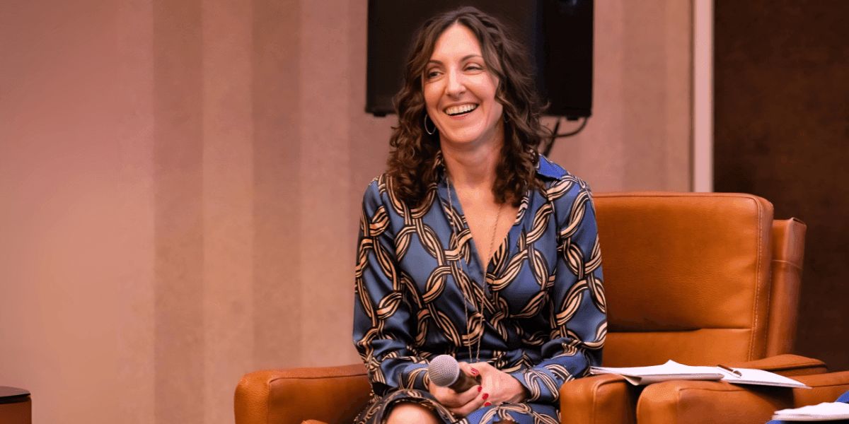 Elizabeth De Sevo smiling while holding a microphone, sitting on a leather chair at an event.