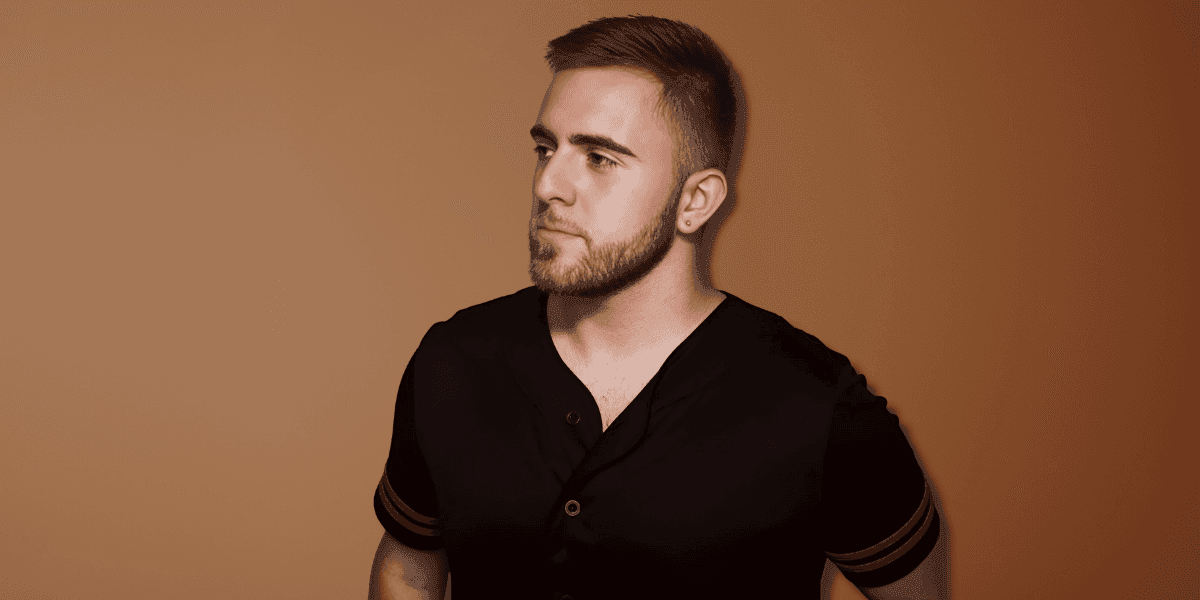 "Portrait of a young man with a short beard, wearing a black shirt with red accents, looking thoughtfully to the side against a warm, neutral background."