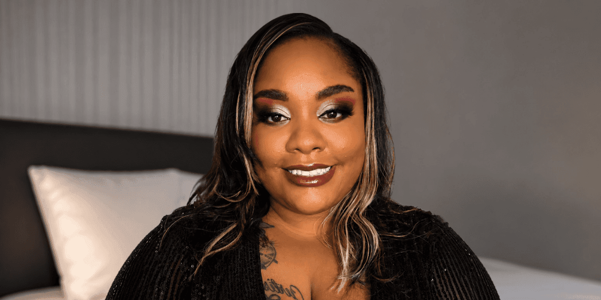 Woman with styled hair and bold makeup smiles in black sequined outfit, seated in softly lit bedroom.