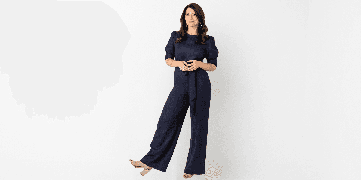 A woman wearing a stylish navy blue jumpsuit with puffed sleeves stands confidently, smiling slightly. She poses with one hand on her waist and the other relaxed at her side, showcasing elegance in front of a clean white background. She completes the look with beige high heels and minimal accessories.