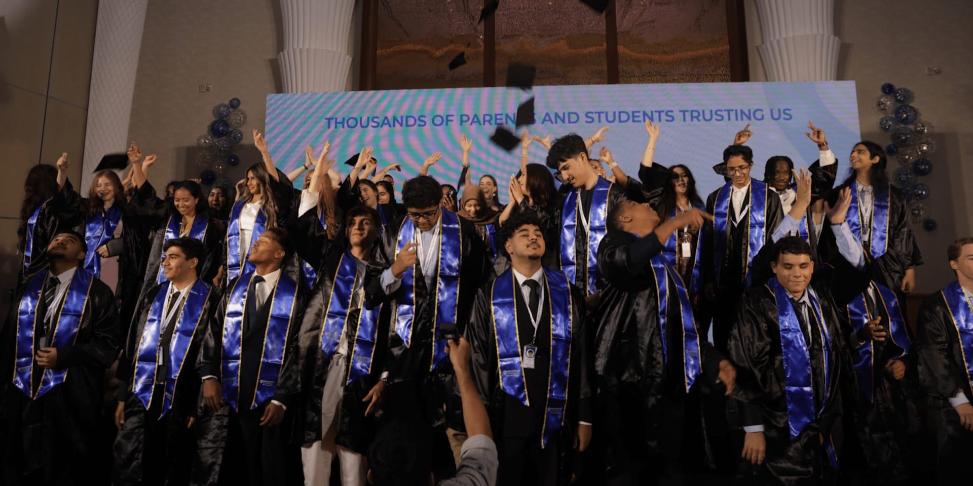 A group of graduates in black caps and gowns with blue stoles are celebrating their graduation, throwing their hats in the air while smiling and cheering. The background shows a banner that reads, "Thousands of Parents and Students Trusting Us.