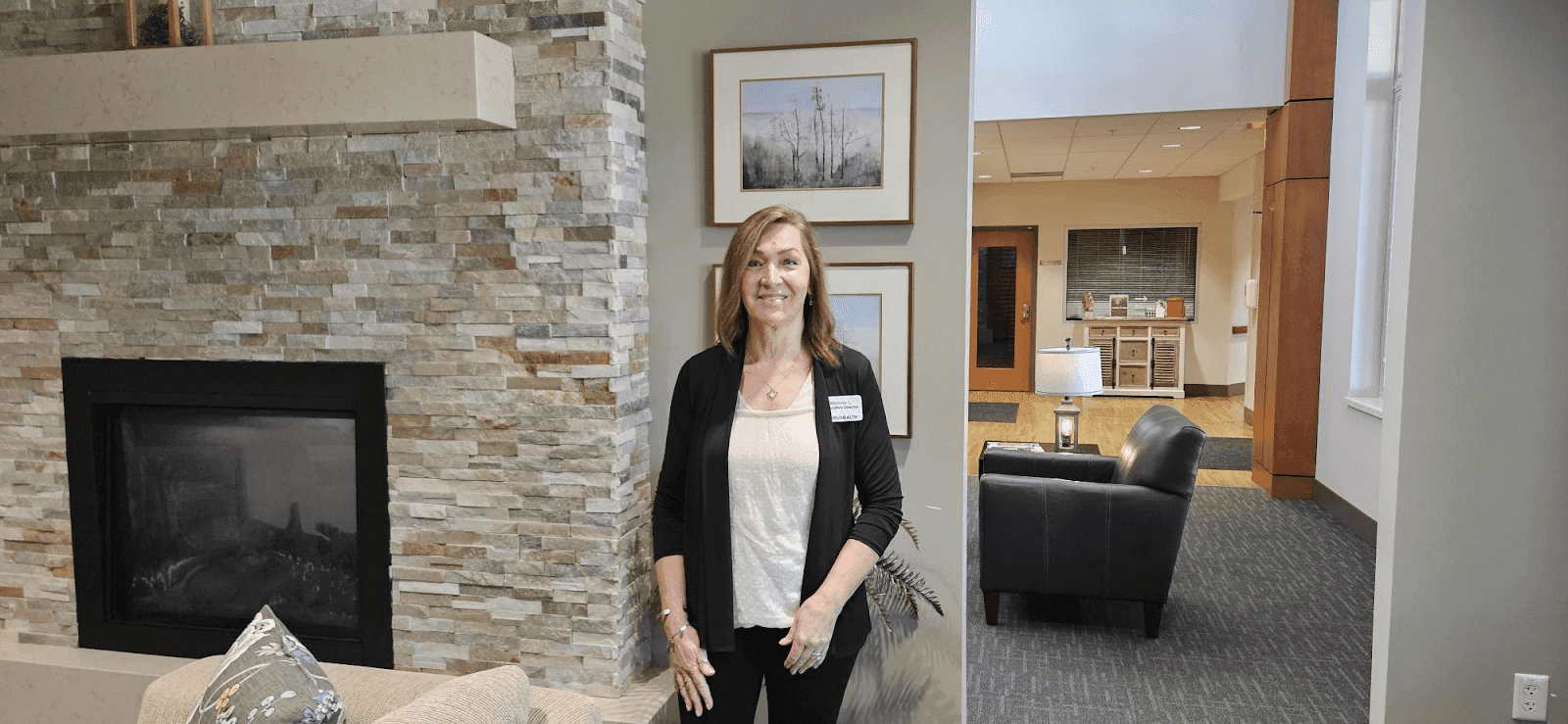 Professional portrait of a woman standing in a modern, well-lit office space with a stone fireplace, wearing a black cardigan and a name tag. The background features stylish furniture and artwork, emphasizing a welcoming and professional atmosphere.